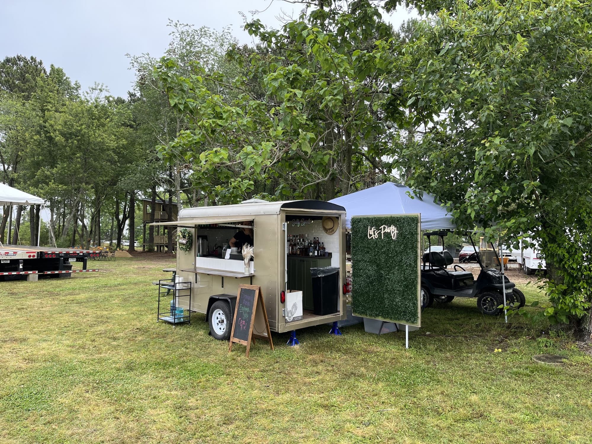 The Blair and its Let's Party greenery wall set up under tall trees at an outdoor field event.