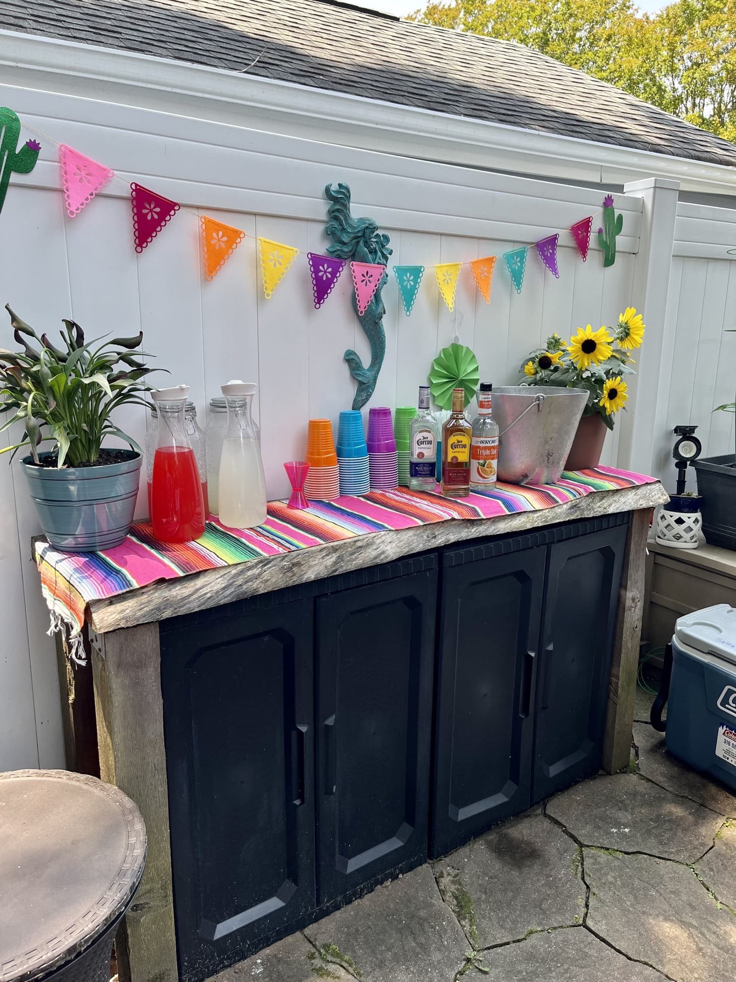 A fiesta-themed margarita station with papel picado, sunflowers and colorful cups on a patio cabinet.