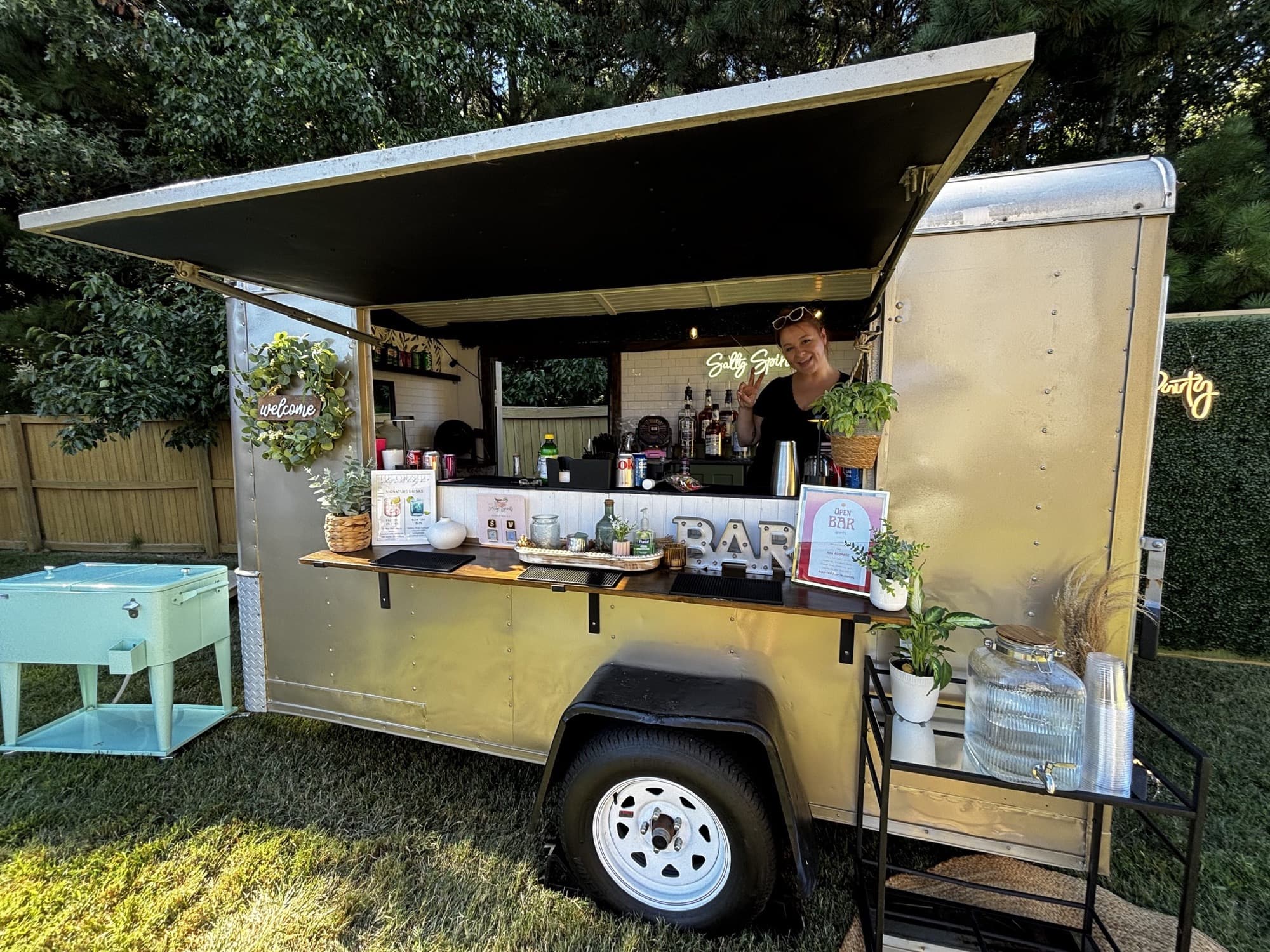 The Blair in golden light at a backyard party, with a bartender in its window, a vintage aqua cooler and the greenery wall beside the trailer.