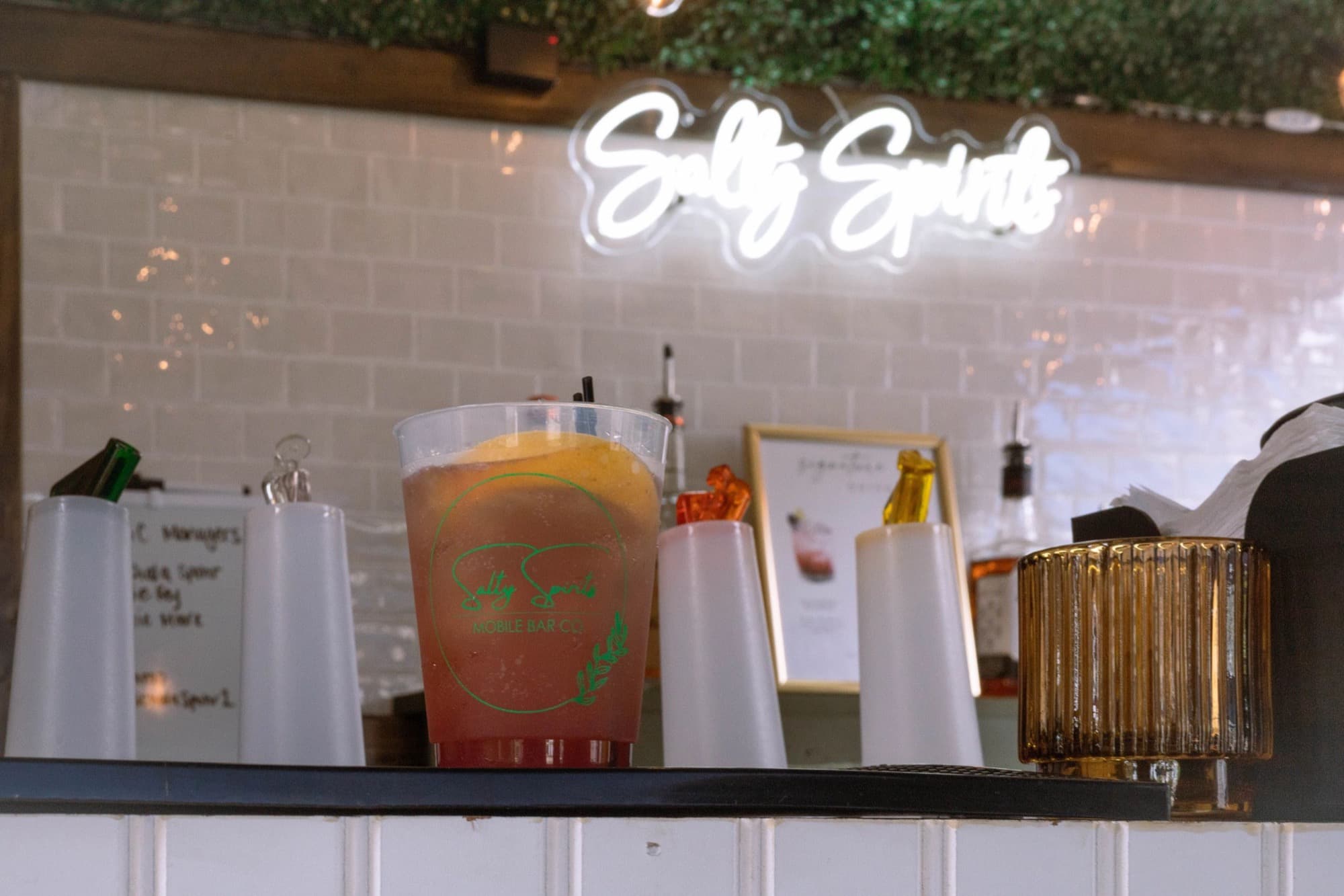 A drink dispenser of punch and a signature drinks frame on the back bar of The Blair, under the white Salty Spirits neon.