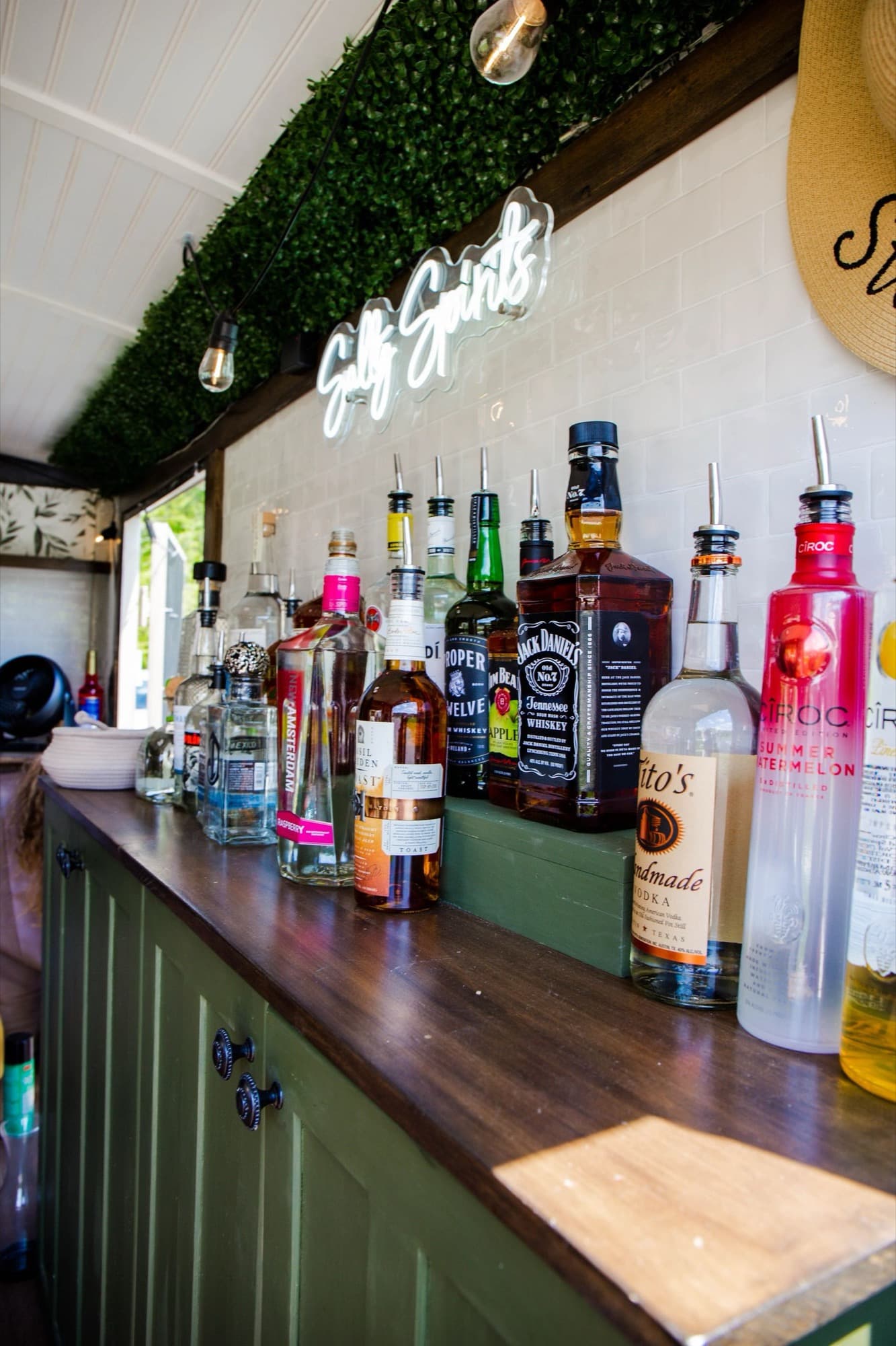 The back bar of The Blair lined with bottles on a wood counter, under the greenery and Salty Spirits neon.