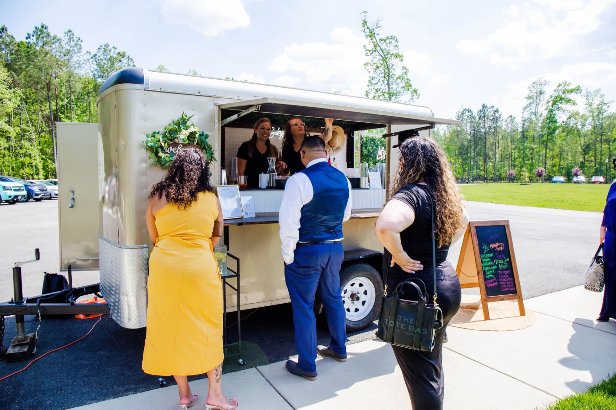 Guests line up at The Blair's service window on a sunny day, with a chalkboard menu by the trailer.